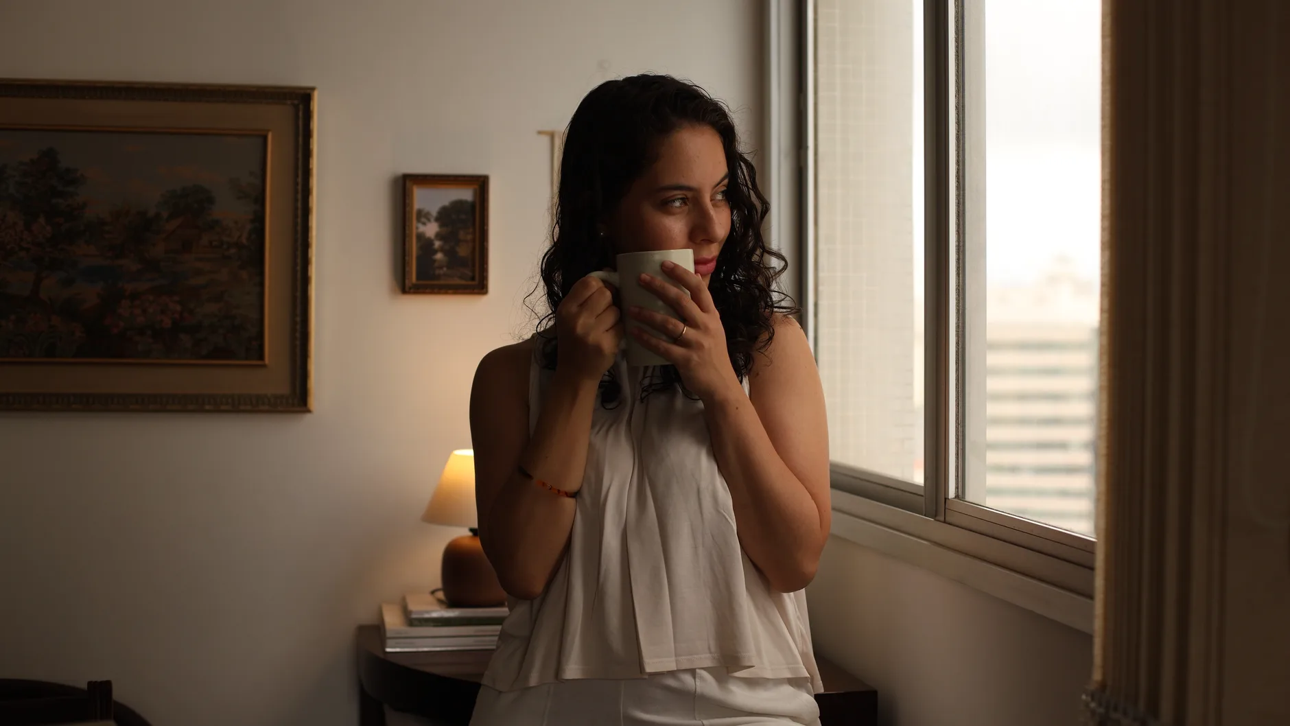 Como criar uma rotina de autocuidado emocional — Jovem mulher contempla a paisagem pela janela enquanto toma café, aprov
